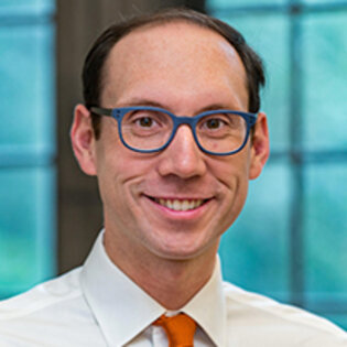 Smiling man with dark hair, blue glasses and light shirt with red tie.