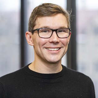 Smiling man with light brown hair, glasses and dark sweater.